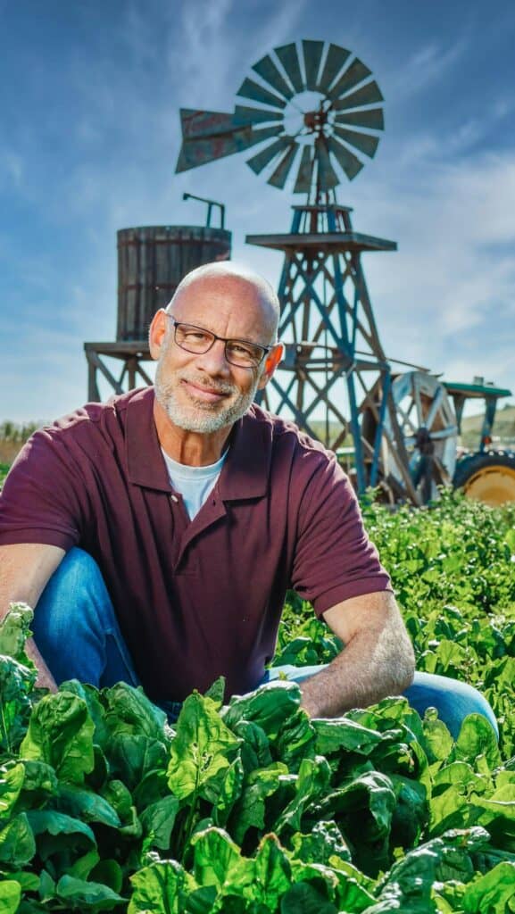 A cheerful bald farmer poses in a lush leafy green field with a vintage windmill in the background.