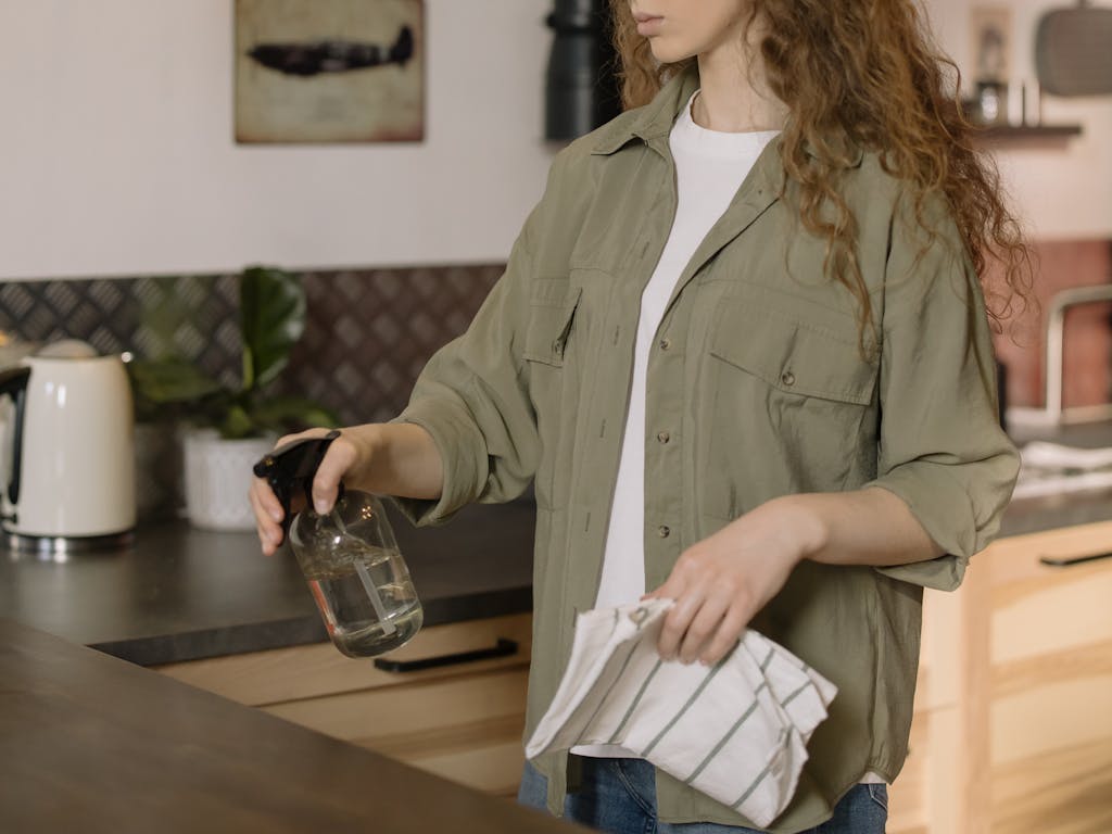 A woman with curly hair cleans a kitchen counter using a spray bottle and cloth.