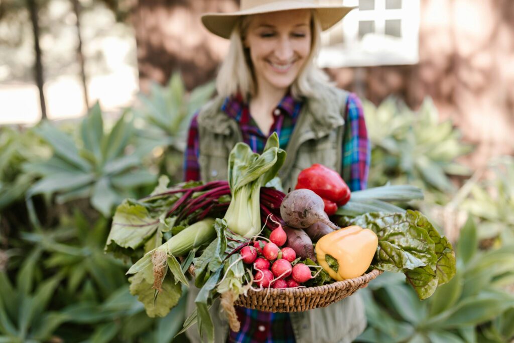 Smiling woman holding fresh farm produce in a basket outdoors, showcasing colorful vegetables.